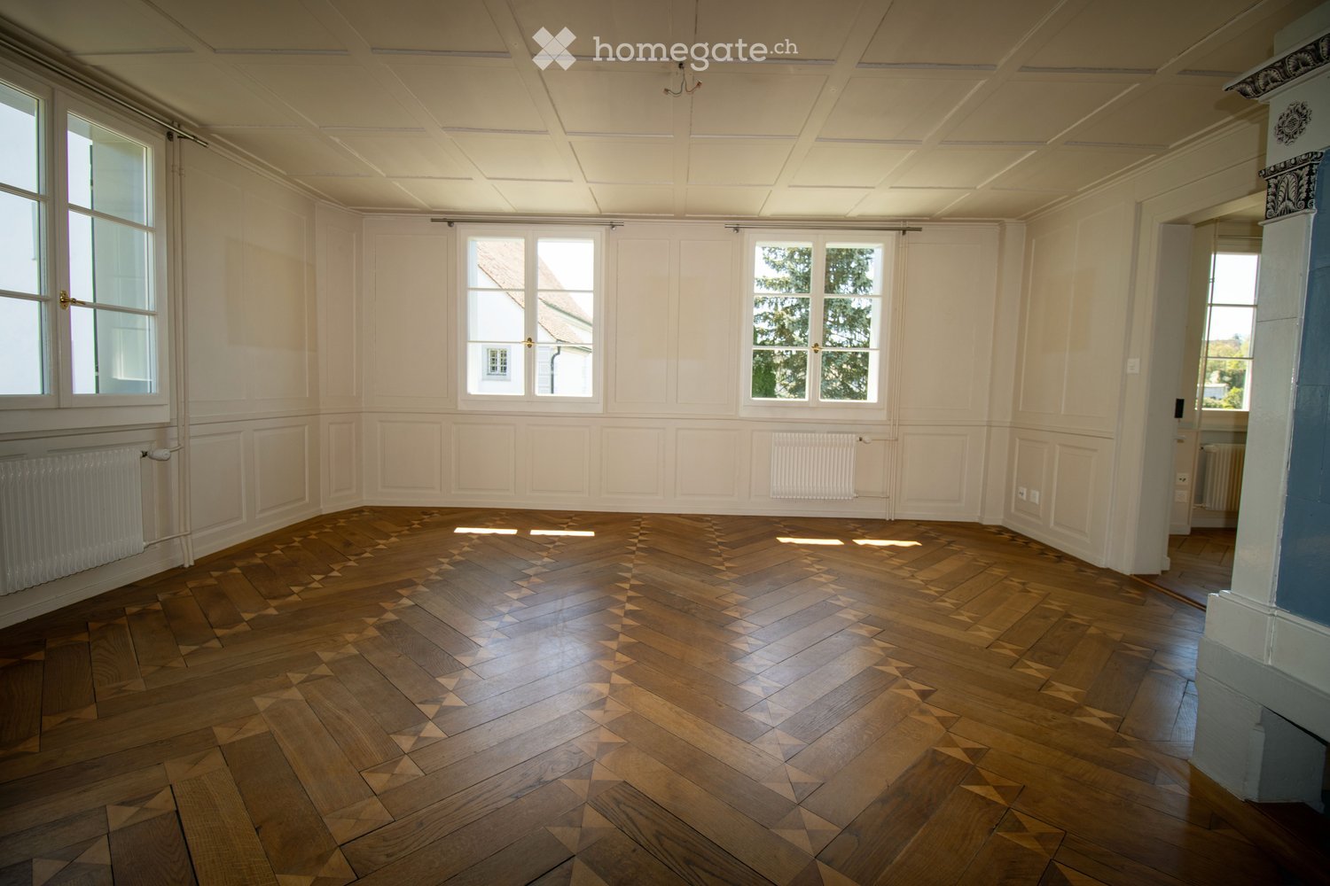 Empty room with wooden herringbone flooring, white paneled walls, large windows, and radiators.