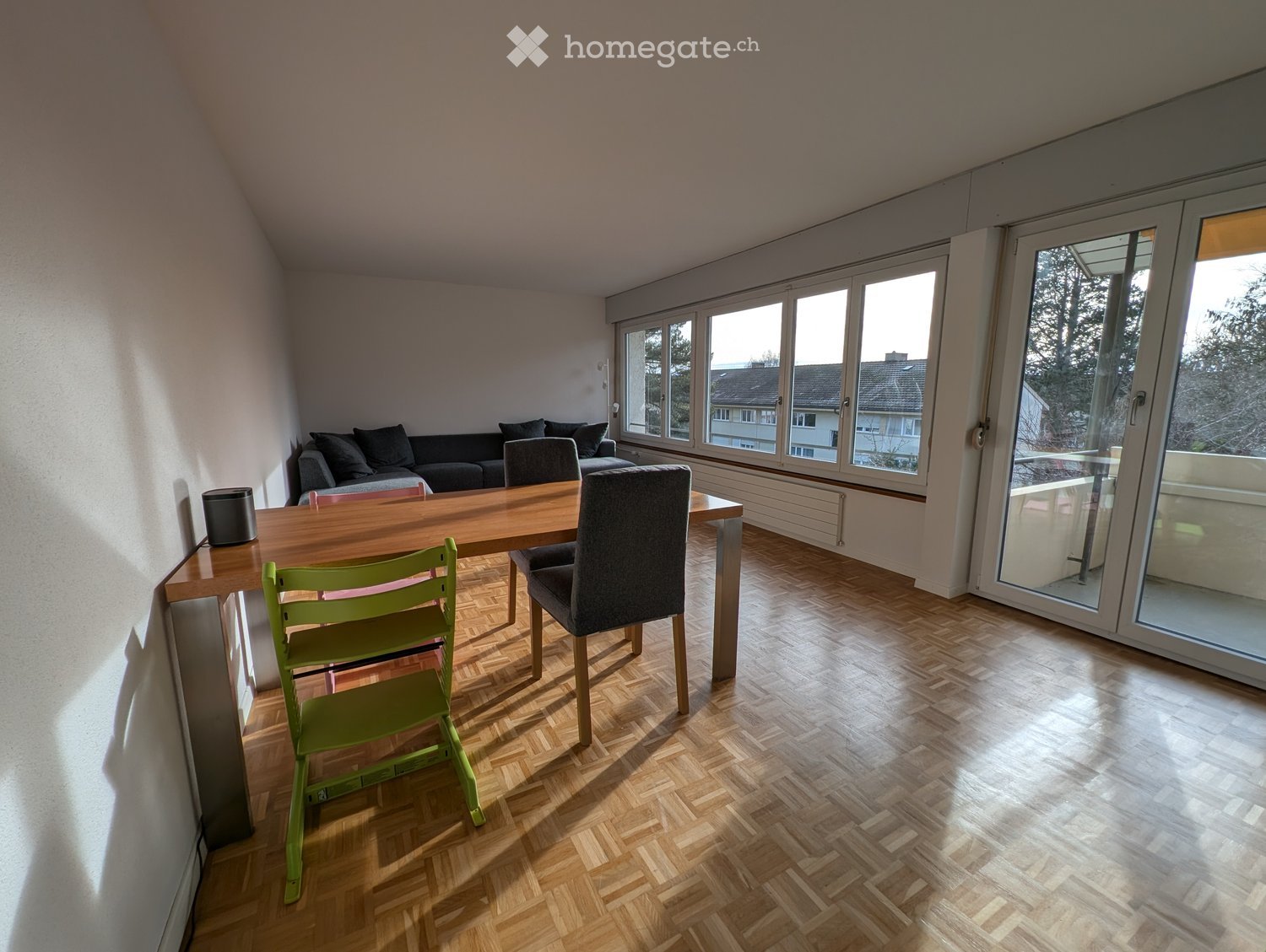 Bright living room with wooden herringbone flooring, large windows, and a sliding glass door leading to a balcony. Features a grey sofa, wooden dining table with green chairs, and a grey armchair.