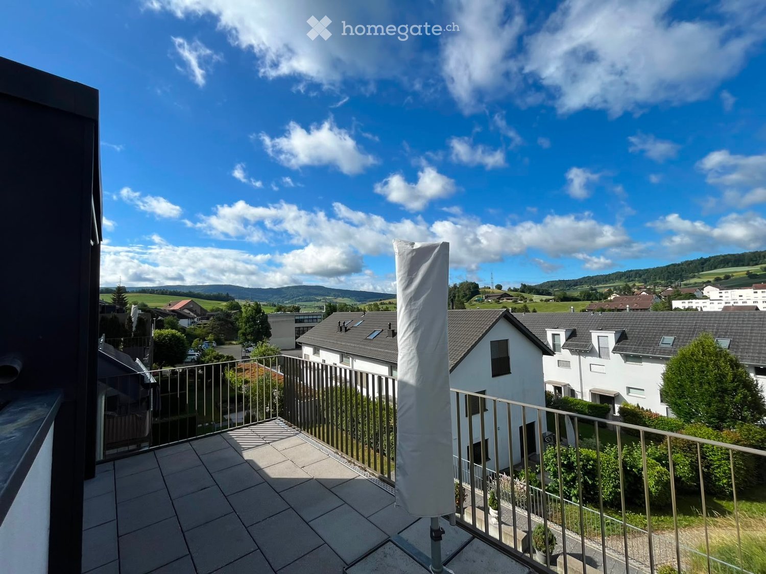 Balcony with tiled floor, metal railing, and a view of a suburban neighborhood with houses, trees, and distant hills under a partly cloudy sky.