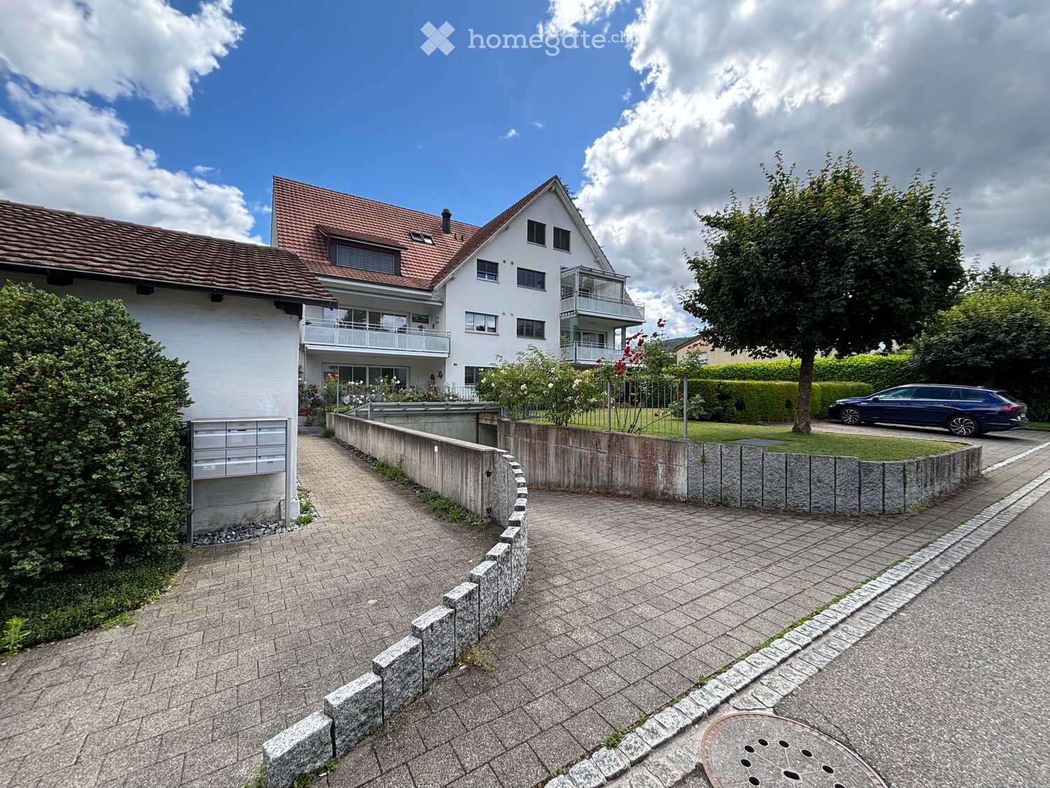 Two-story house with white exterior, red-tiled roof, and a balcony on the upper floor. The house is surrounded by a low stone wall and a paved pathway leading to the entrance. There is a mailbox on the left side and a blue car parked on the right.