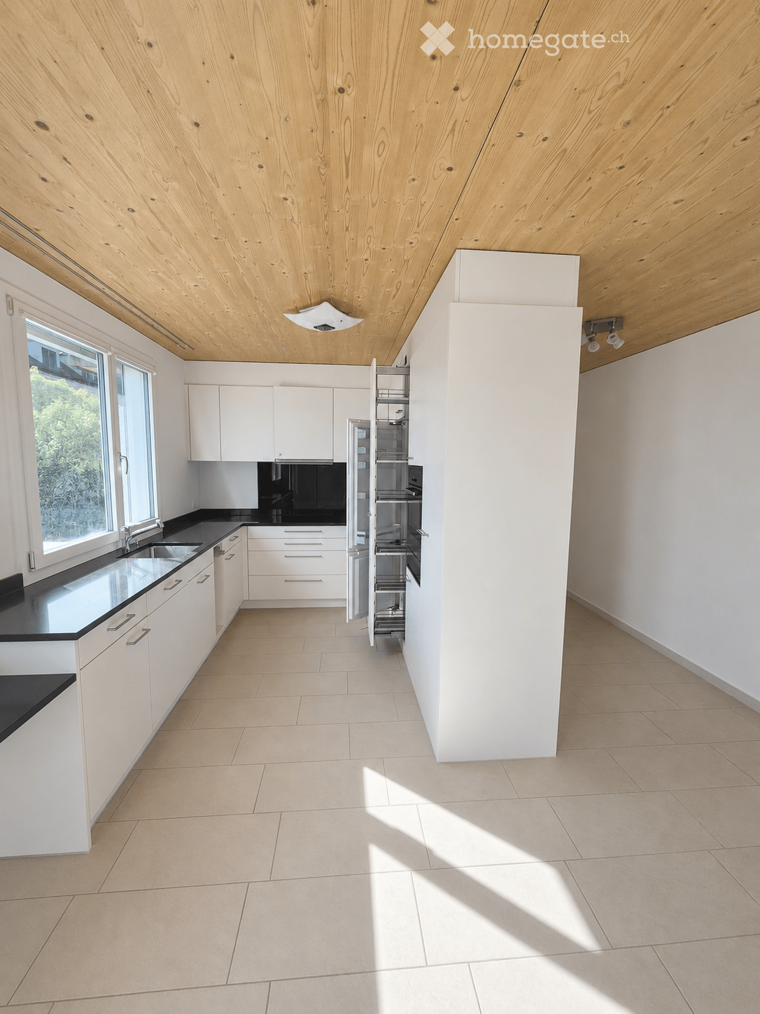 Modern, minimalist kitchen with white cabinets, black countertops, and a stainless steel sink. The space features a sleek, open design with a tall pantry unit and ample natural light from large windows.