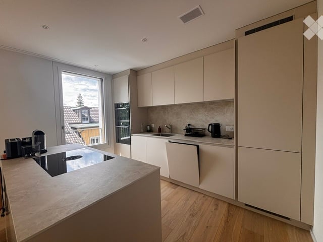 Modern kitchen with light-colored cabinets, a central island, and a large window offering a view of the outside. Appliances include a fridge, oven, and coffee maker.