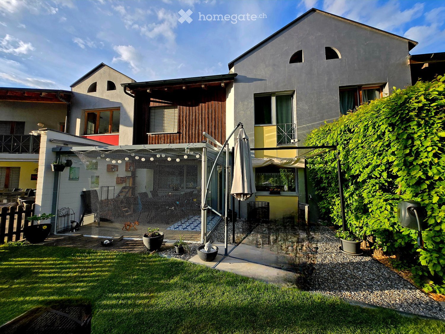 Garden / Outdoor Area. A well-maintained garden with a lawn, gravel pathways, potted plants, and a covered patio area with an umbrella and outdoor furniture.