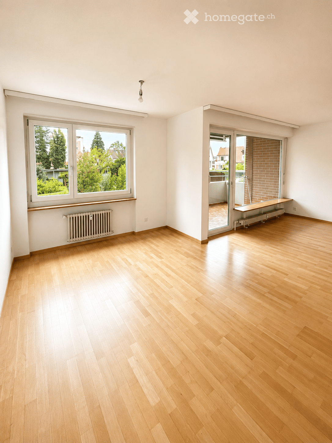 Empty room with light wooden flooring, white walls, large windows, and a sliding glass door leading to a balcony area.