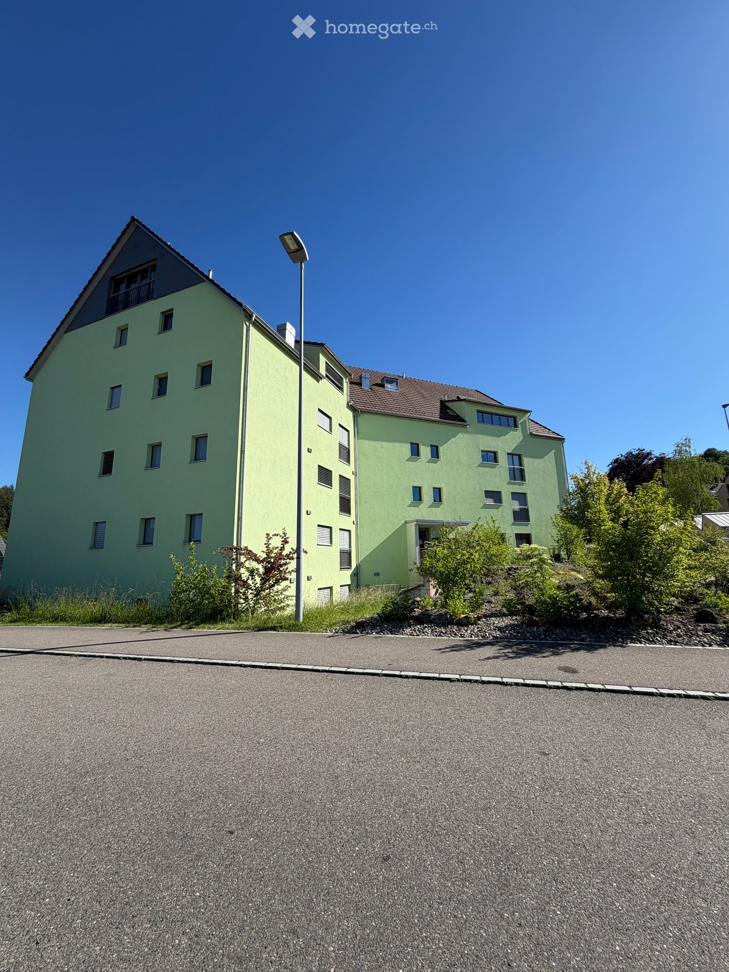 Two-story green building with multiple windows and a balcony on the upper floor, surrounded by greenery and a paved area.