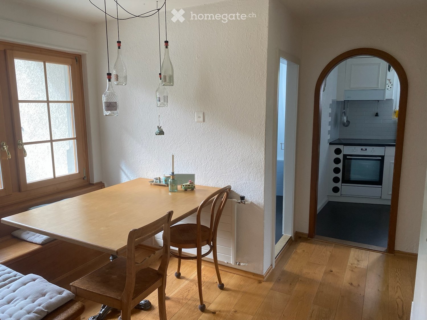 Bright dining area with wooden table and chairs, a bench seat, and a view into a modern kitchen with an oven and hanging pendant lights.