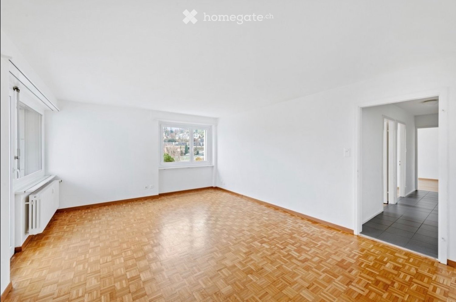 Empty room with white walls, wooden herringbone flooring, and large windows offering a view of greenery. Radiator visible under one window, and an open doorway leads to another room with tiled flooring.