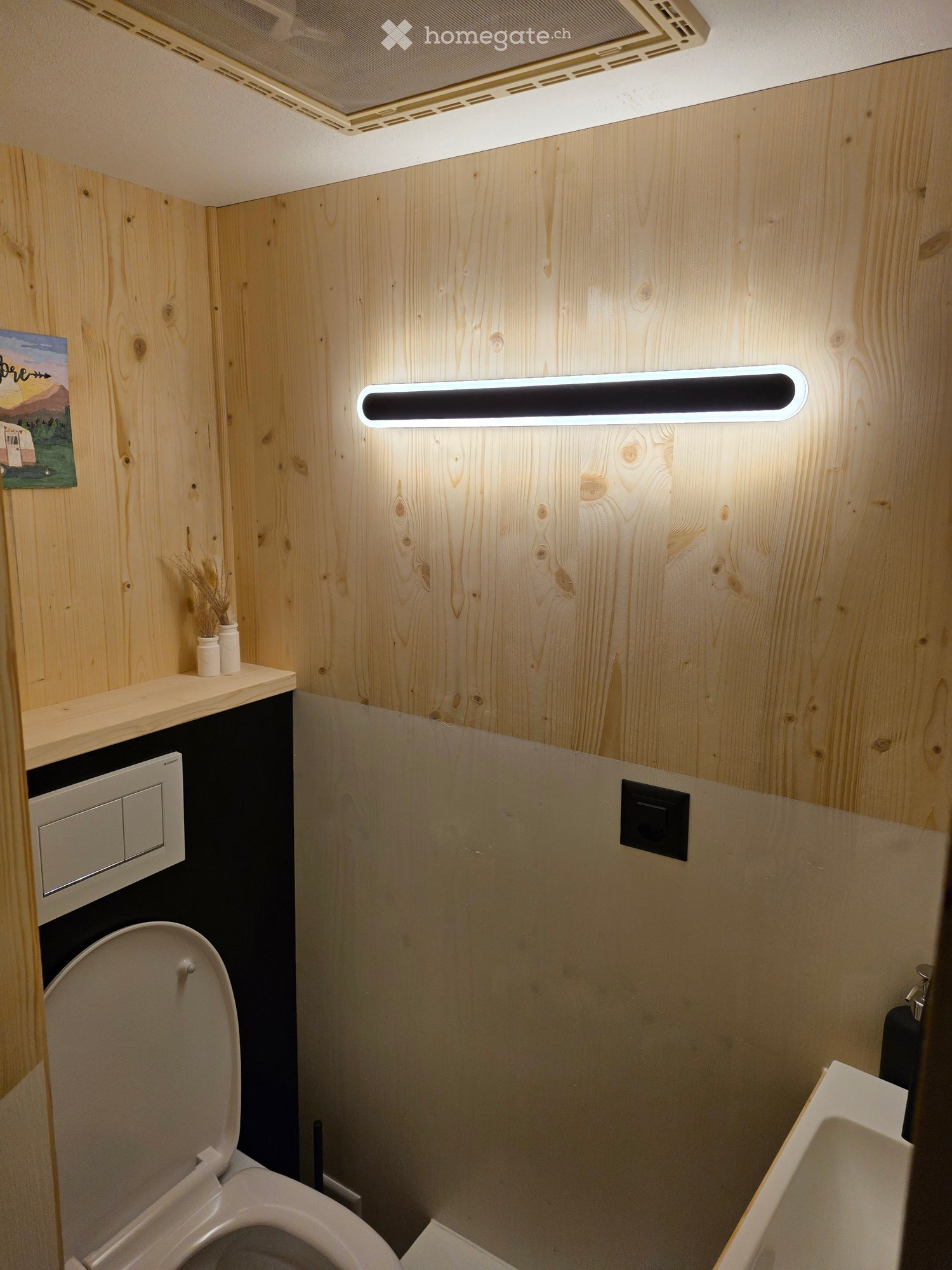 Compact bathroom with wooden walls, a toilet, a sink, a small shelf with vases, and modern lighting fixtures.