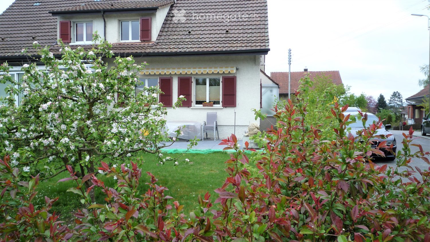 Two-story house with red shutters, tiled roof, and a small garden with flowering trees and shrubs. A car is parked on the street.