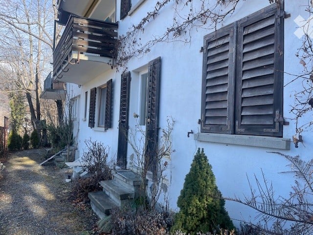 Two-story white building with wooden shutters, a balcony on the upper floor, and a small garden area with steps leading up to the entrance.