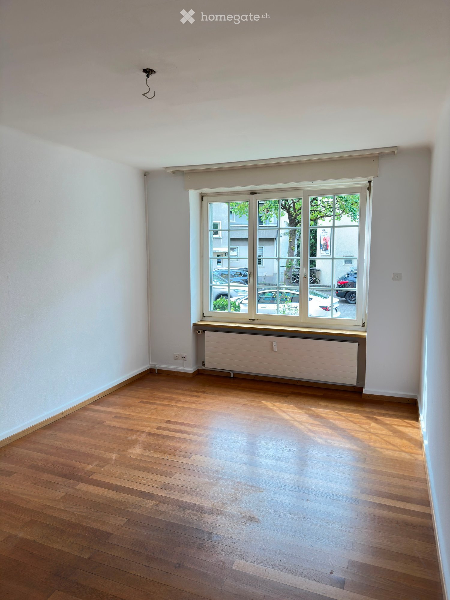 Empty room with wooden flooring, white walls, large window with blinds, radiator below the window, and visible electrical outlets.