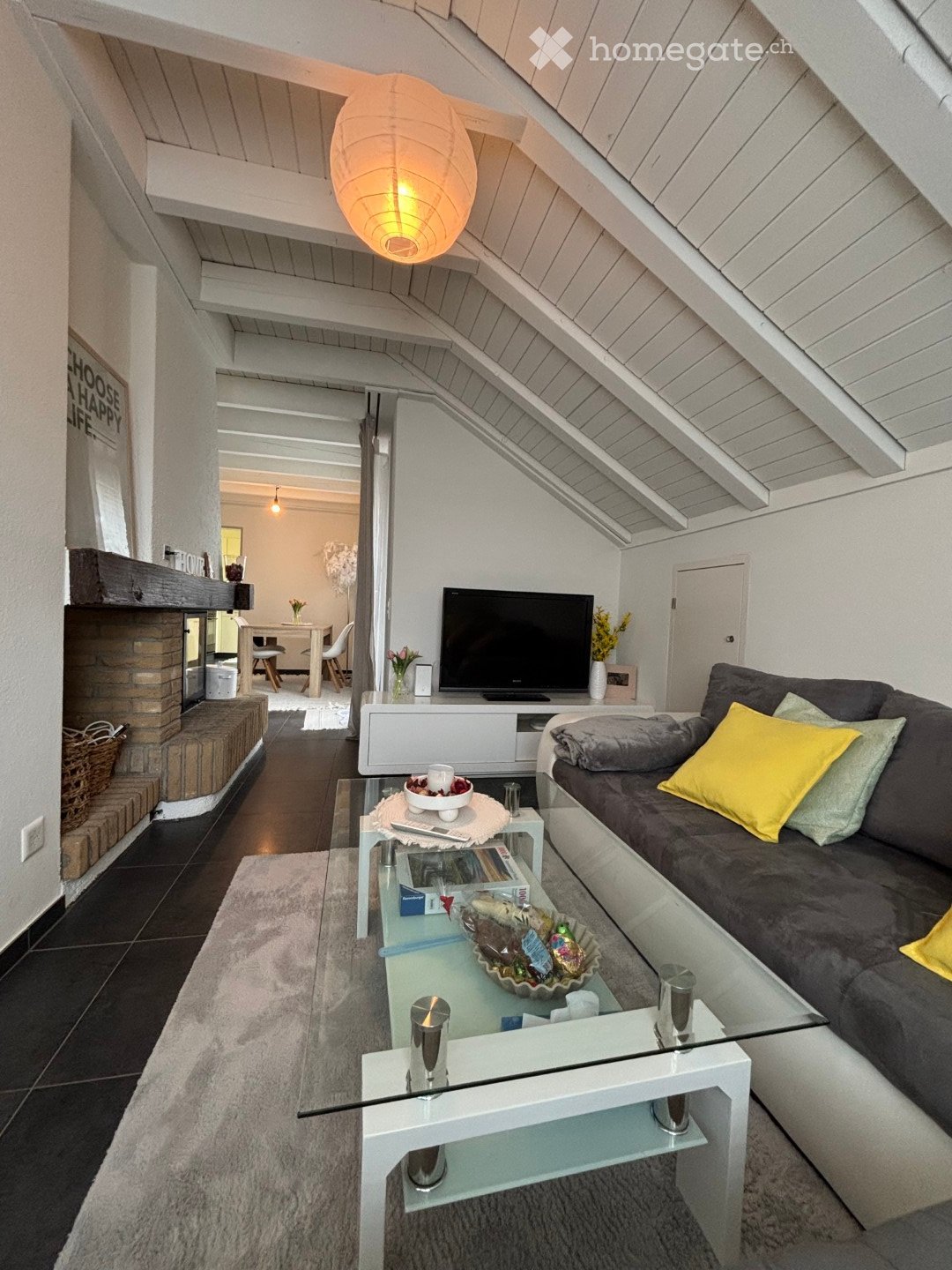 Modern living room with a slanted wooden ceiling, a grey sofa with colorful cushions, a glass coffee table, a fireplace, and a television on a white stand. The room features a cozy rug and a small dining area visible in the background.