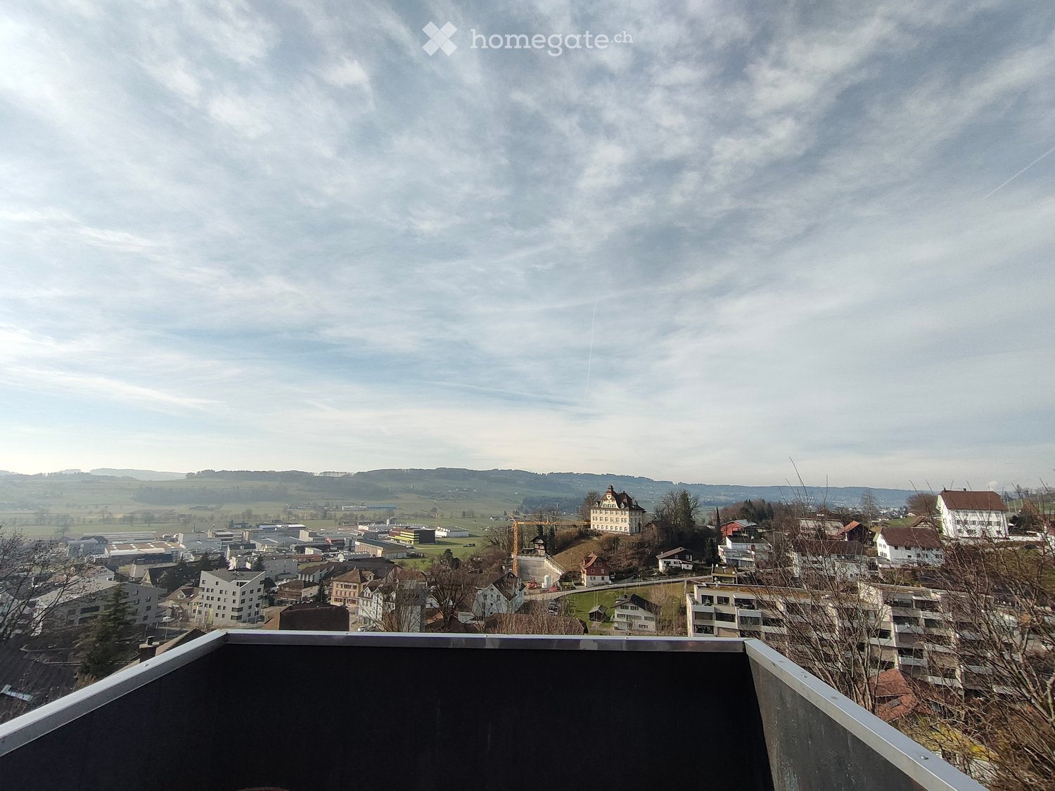 View from a balcony or terrace overlooking a town with buildings, a castle, and surrounding countryside under a partly cloudy sky.