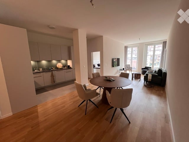 Open-plan living and dining area with light wood flooring, modern grey kitchen cabinets, a round dining table with four chairs, a black sofa, and a television mounted on the wall.