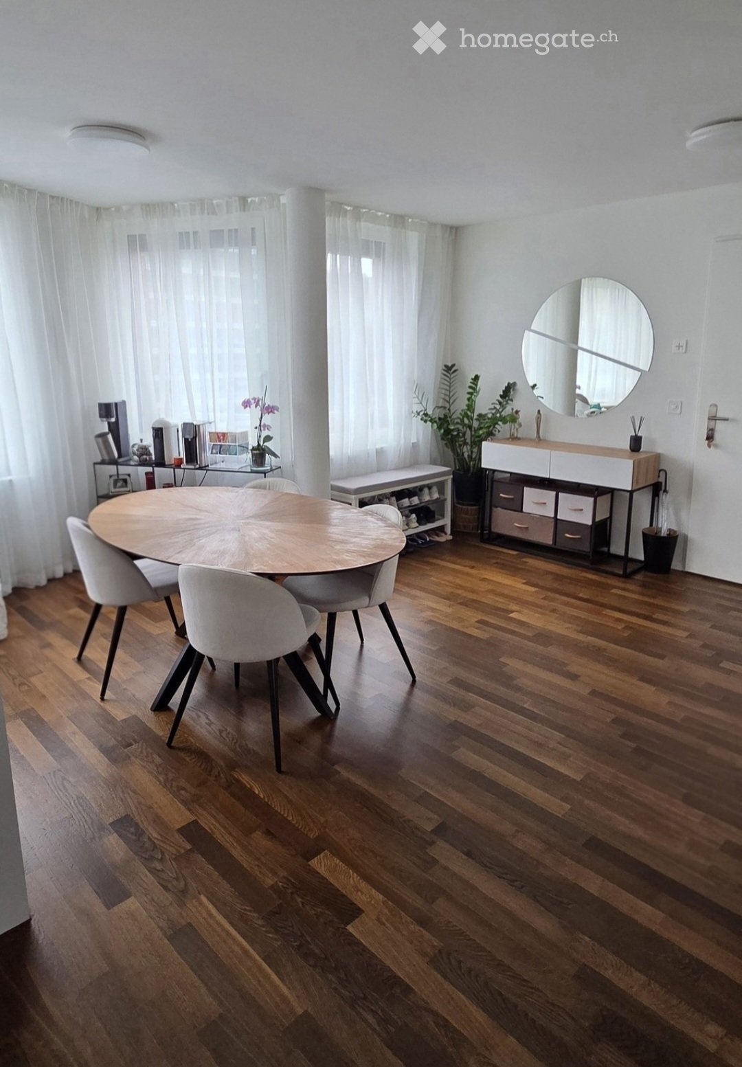 Bright dining area with wooden floor, round wooden table surrounded by white chairs, storage unit with drawers, bench, potted plants, and a round mirror on the wall.