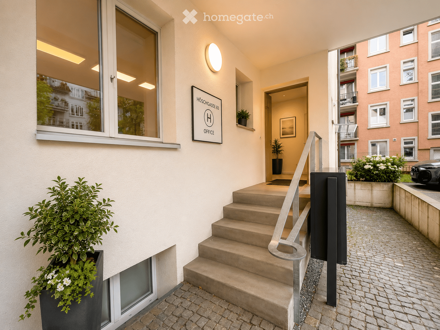 Entrance area with a staircase leading upwards, featuring a modern railing, potted plants, and a sign reading "HÖSCHGASSE 83 OFFICE".