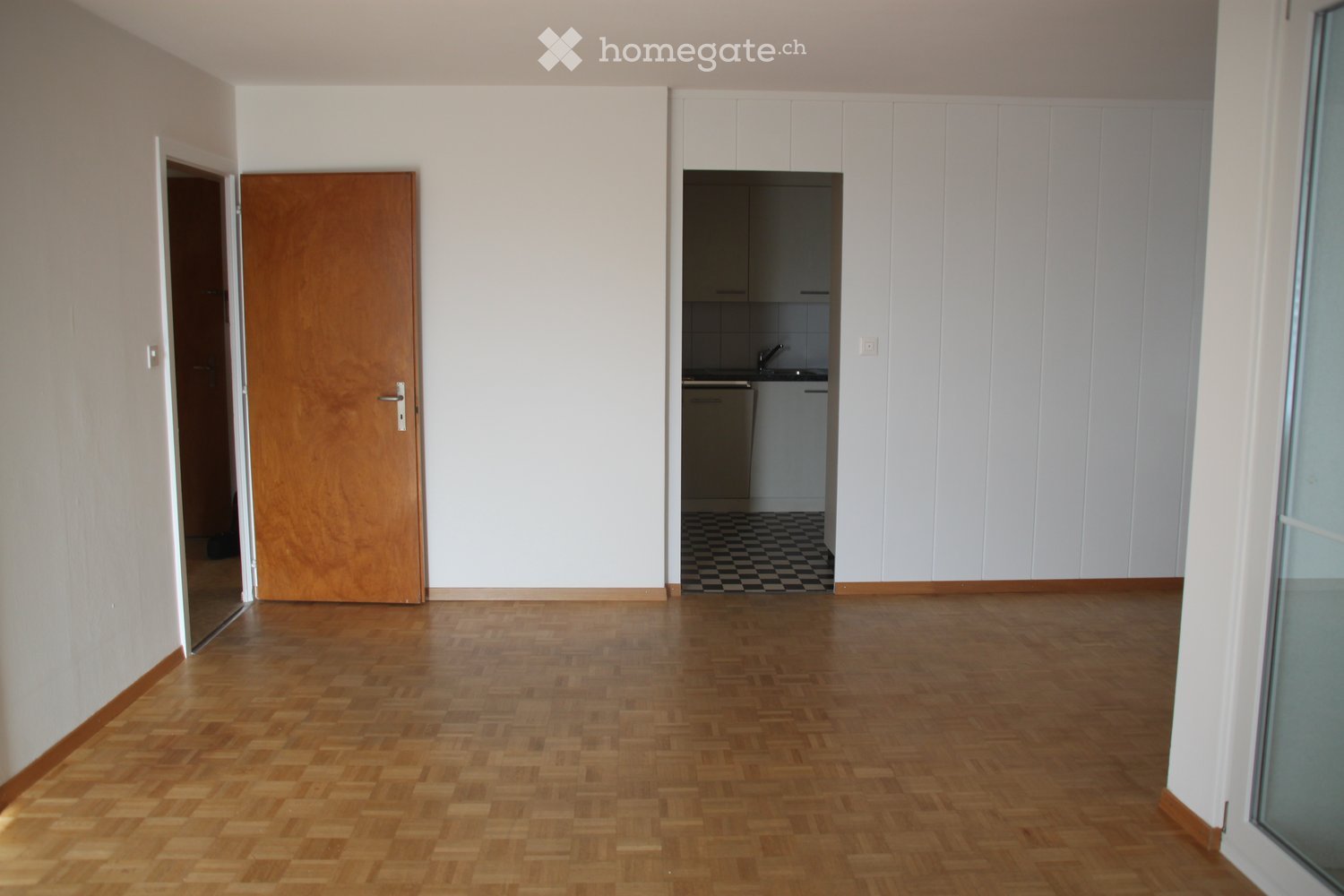 Empty room with wooden flooring, white walls, and a doorway leading to a small kitchen area with white cabinets and a checkered floor.