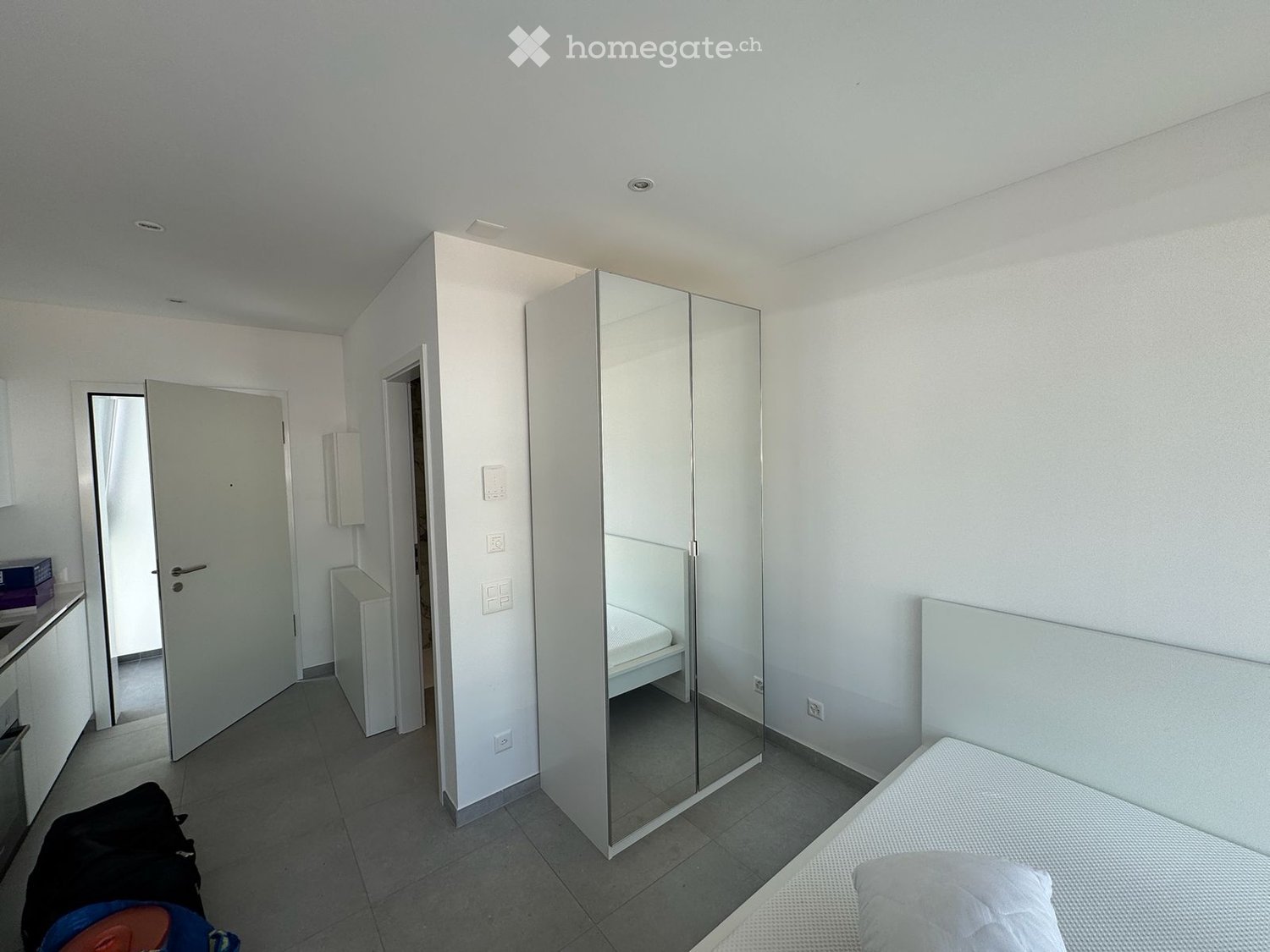 Modern, minimalist bedroom with a single bed, large mirrored wardrobe, and tiled floor. The room features white walls and a neutral color palette.