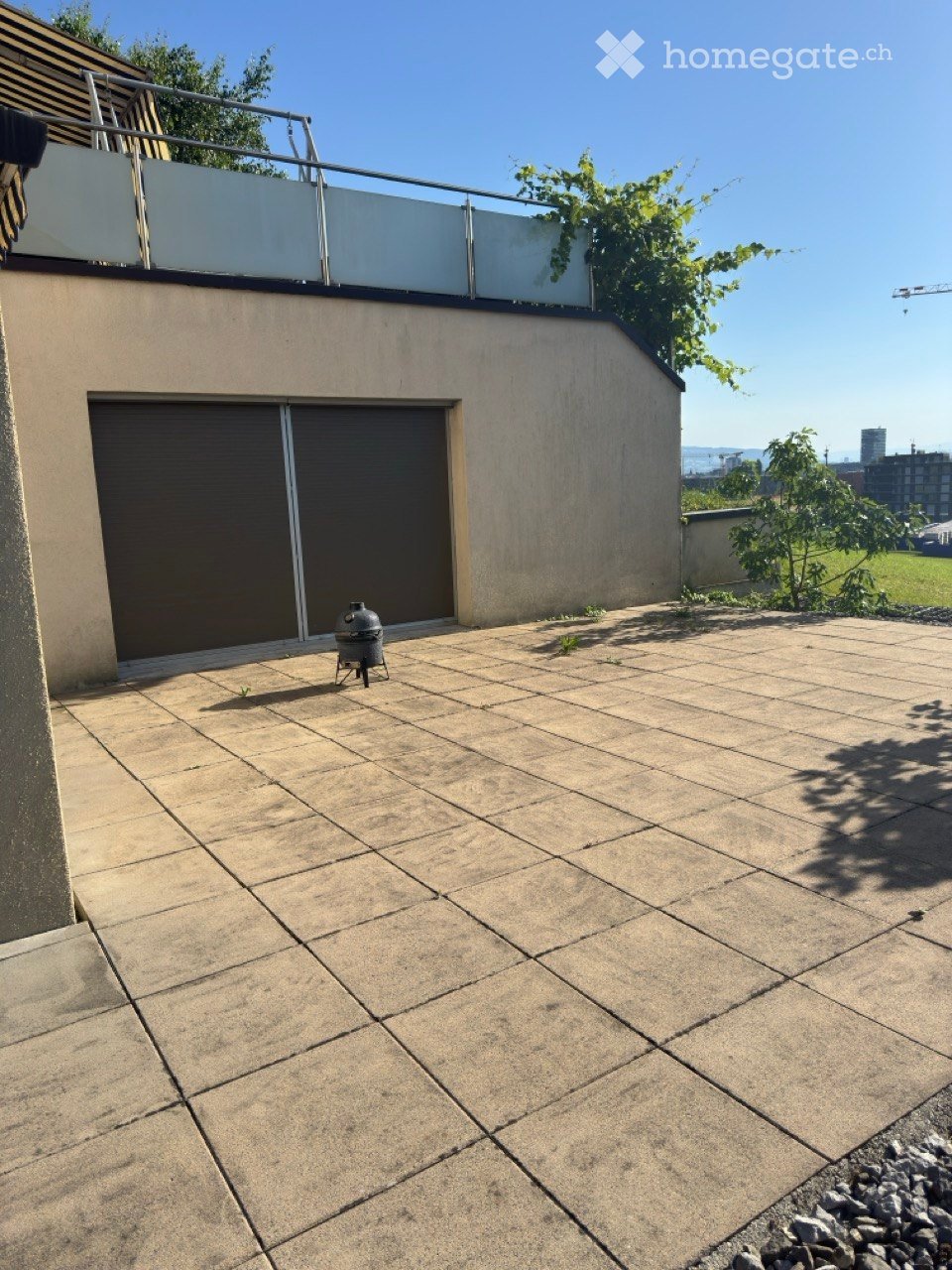 A modern rooftop terrace with a tiled concrete floor, a small barbecue, and a view of the city skyline and water in the distance.