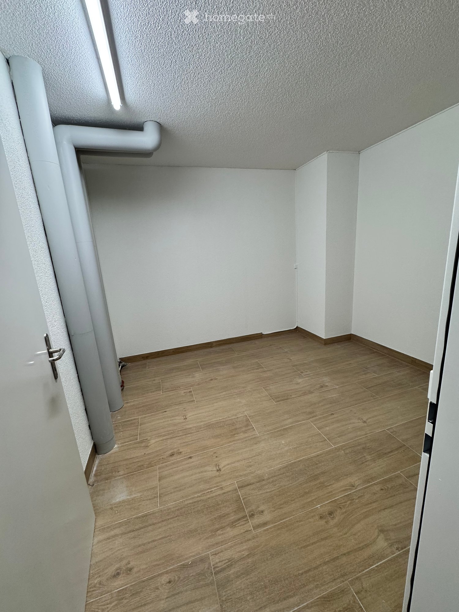 Empty room with light wooden flooring and white walls, featuring a door and a visible pipe in the corner.