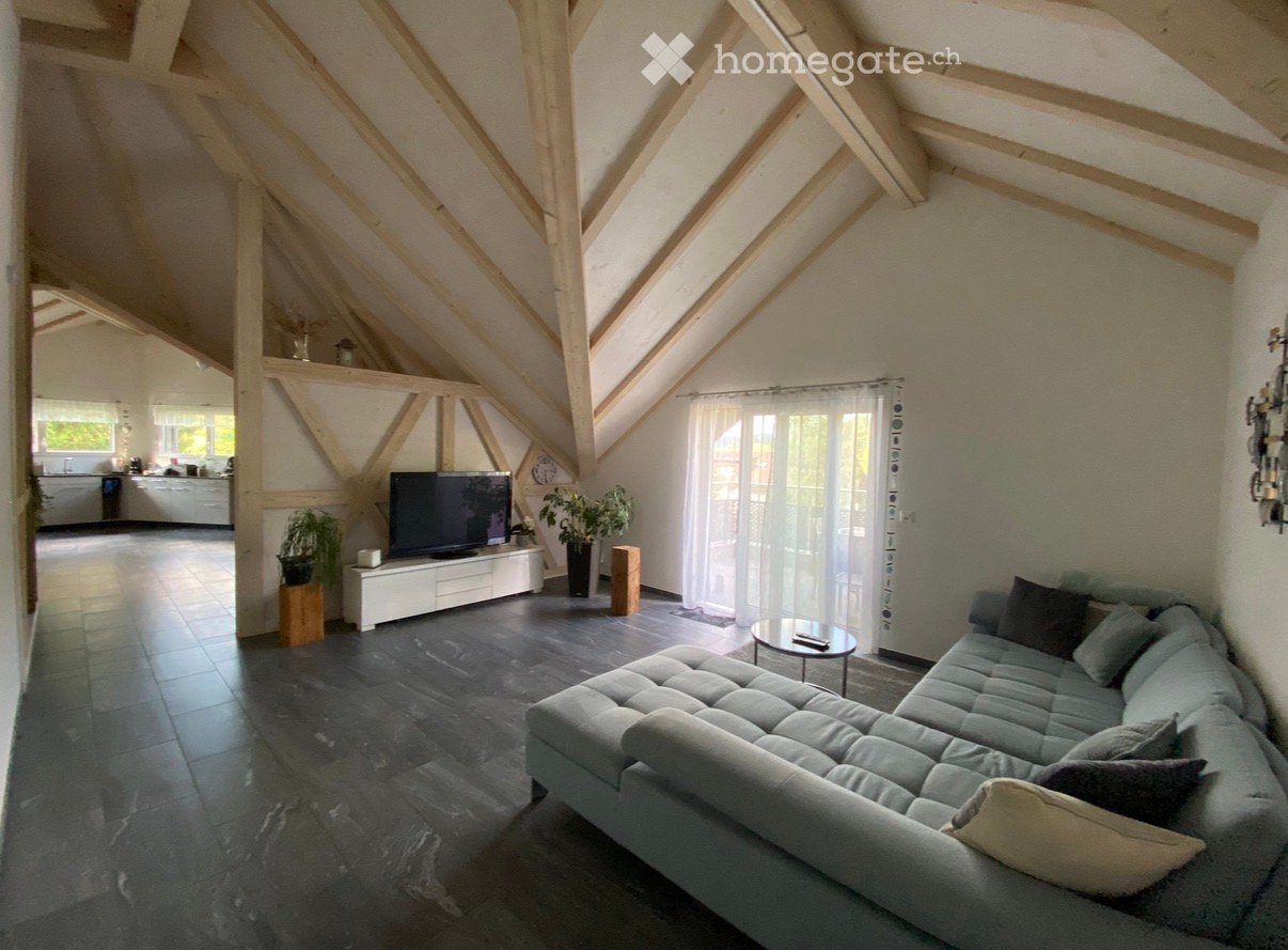 Modern living room with exposed wooden beams, grey sectional sofa, coffee table, flat-screen TV on a white cabinet, and plants. The room features large windows with sheer curtains and a dark tiled floor.