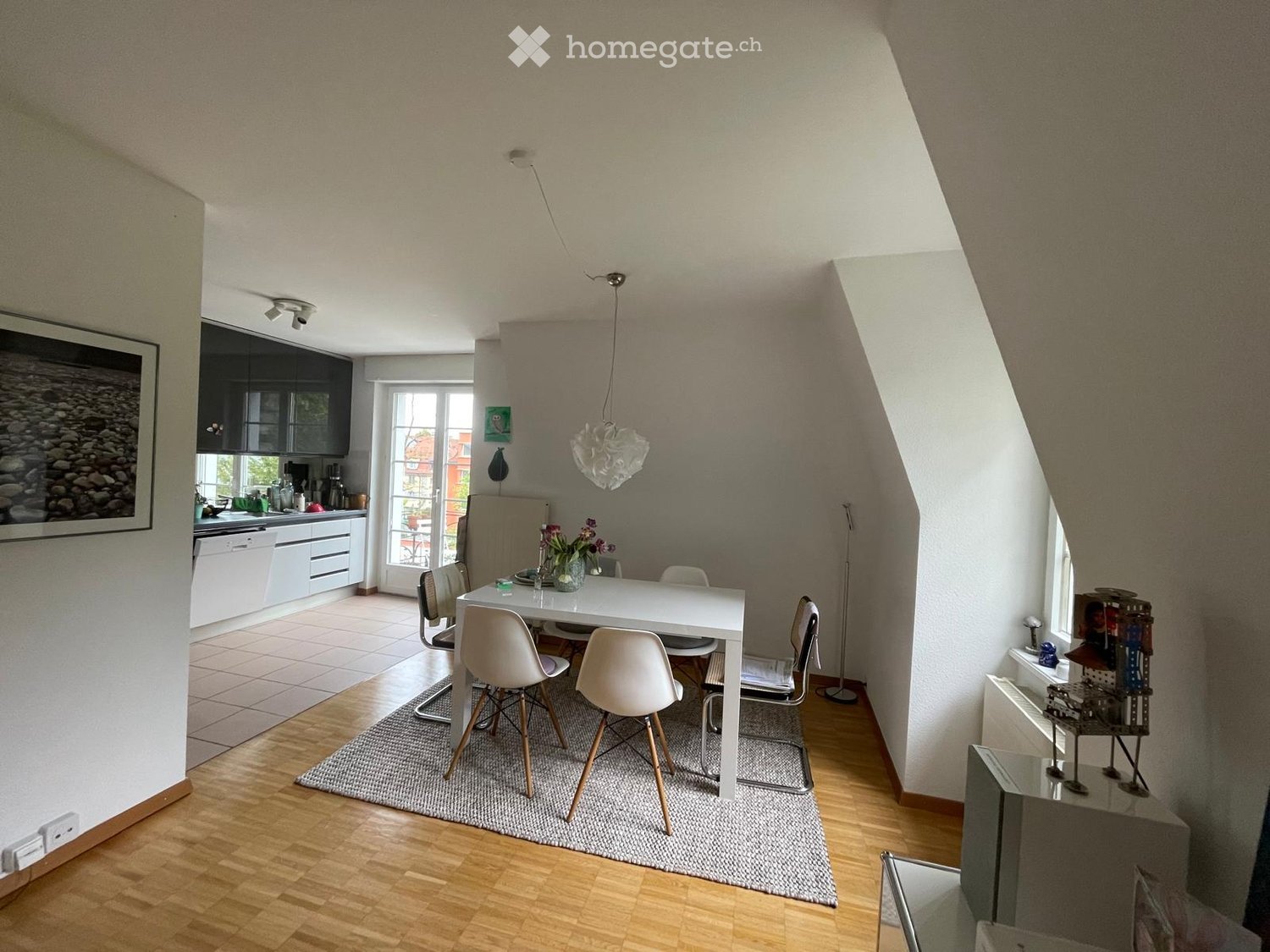 Bright, open-plan living and dining area with modern furniture, including a white dining table with chairs, a kitchen area with cabinets and appliances, and decorative elements like a framed picture and a vase with flowers.