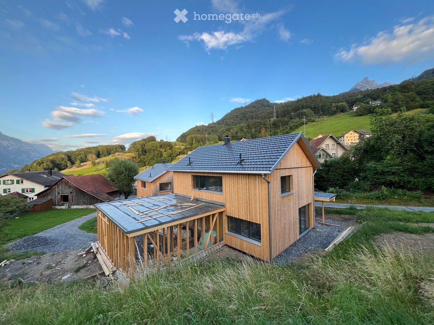 Modern wooden house with grey roof, surrounded by green landscape and mountains.