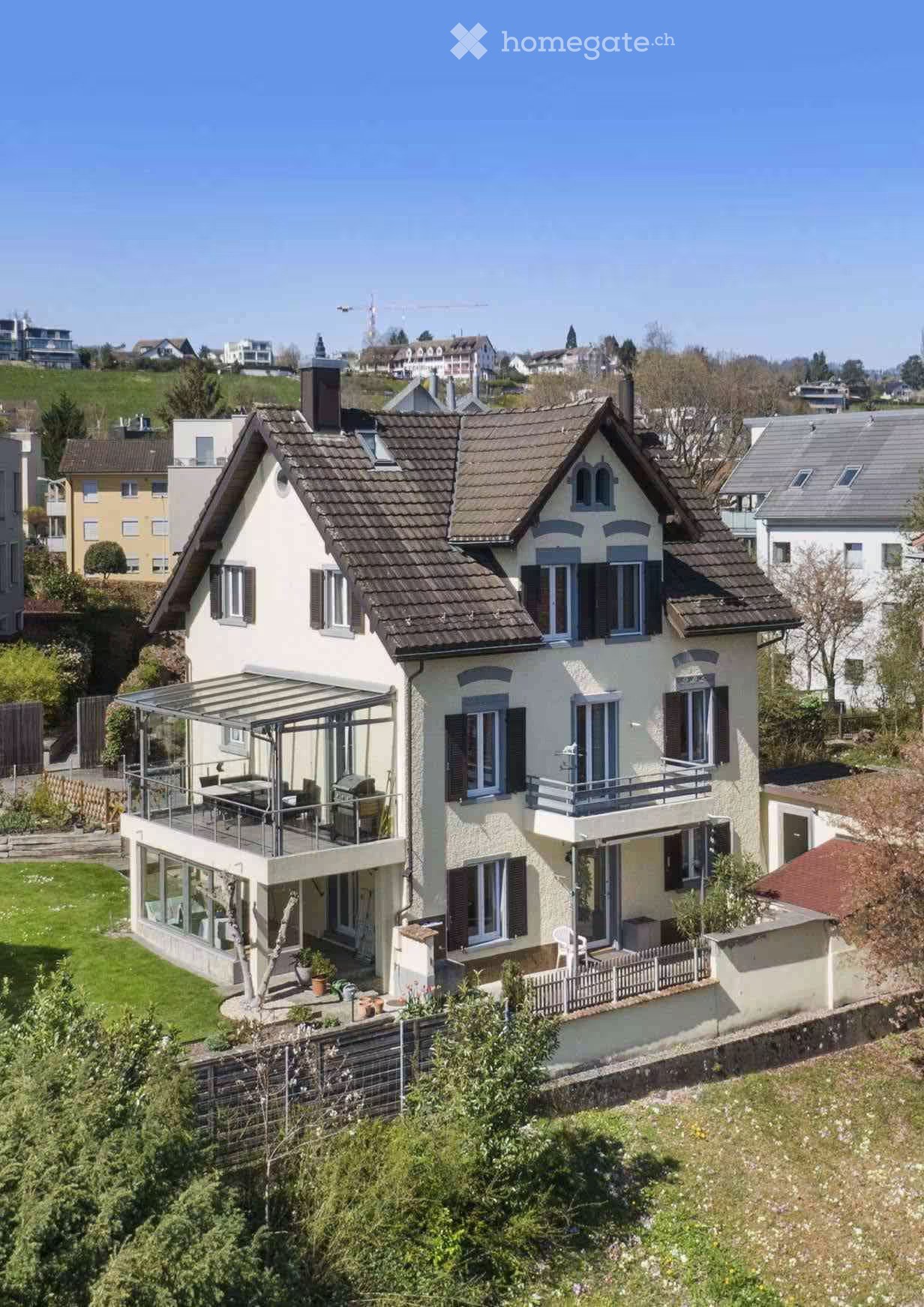 Two-story house with a tiled roof, multiple windows, and a balcony. The house features a covered patio area with outdoor furniture and a small garden with potted plants.