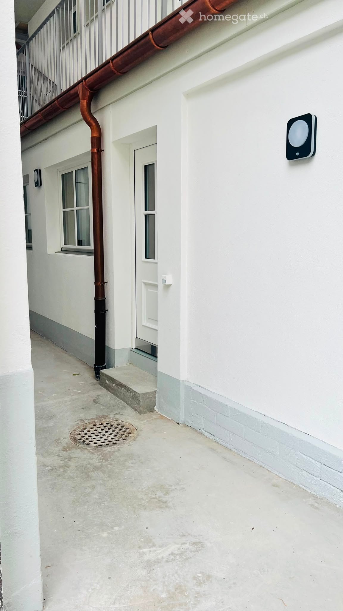 A narrow concrete pathway next to a white building with a small door, a metal grate, and a balcony above.