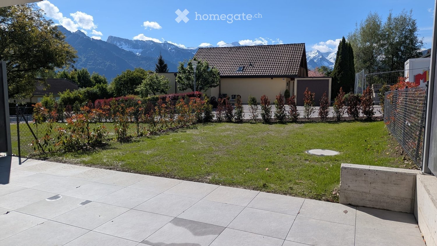 Well-maintained garden with green lawn, trimmed bushes, and a small house in the background, with mountain views.