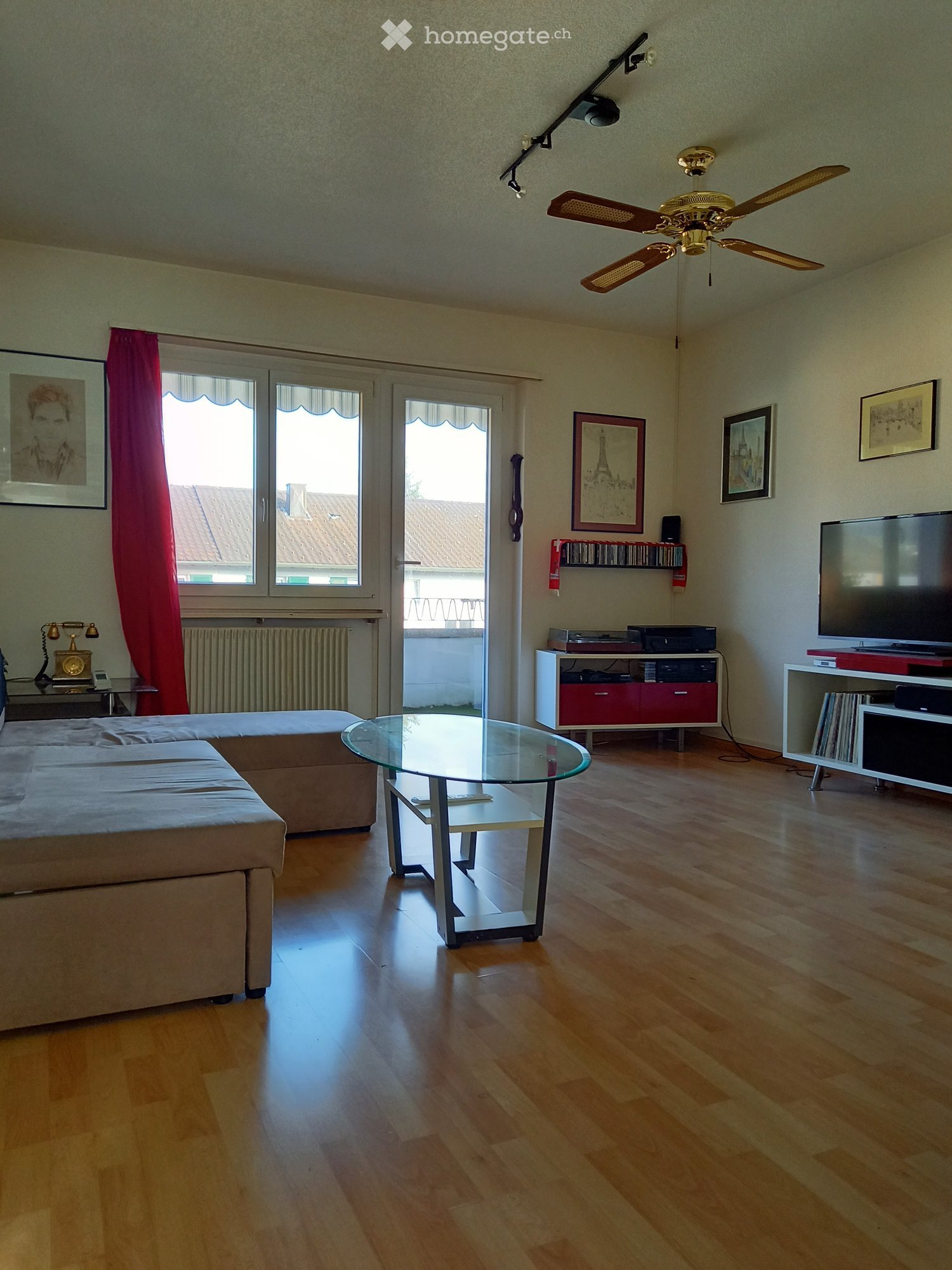 Bright living room with wooden flooring, large windows, a sofa, a glass coffee table, TV stand, and a ceiling fan.