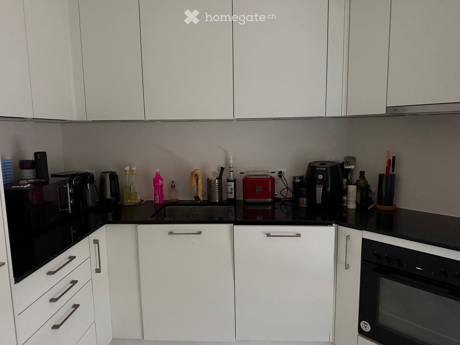 Modern kitchen with white cabinets, black countertops, and various appliances including an oven, toaster, coffee maker, and fridge. Several bottles and cups are placed on the counter.