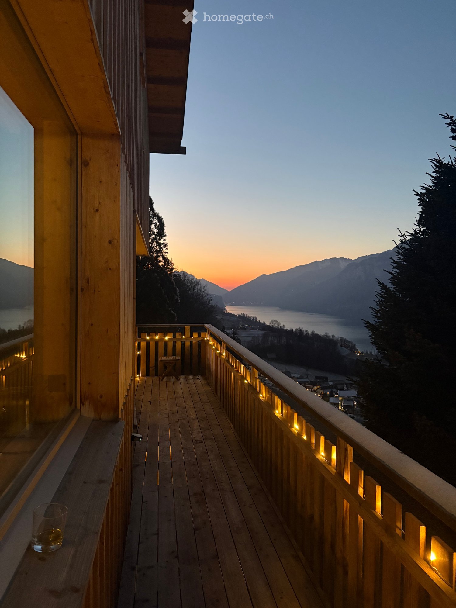 Wooden balcony with railing lights, overlooking a lake and mountains at sunset.
