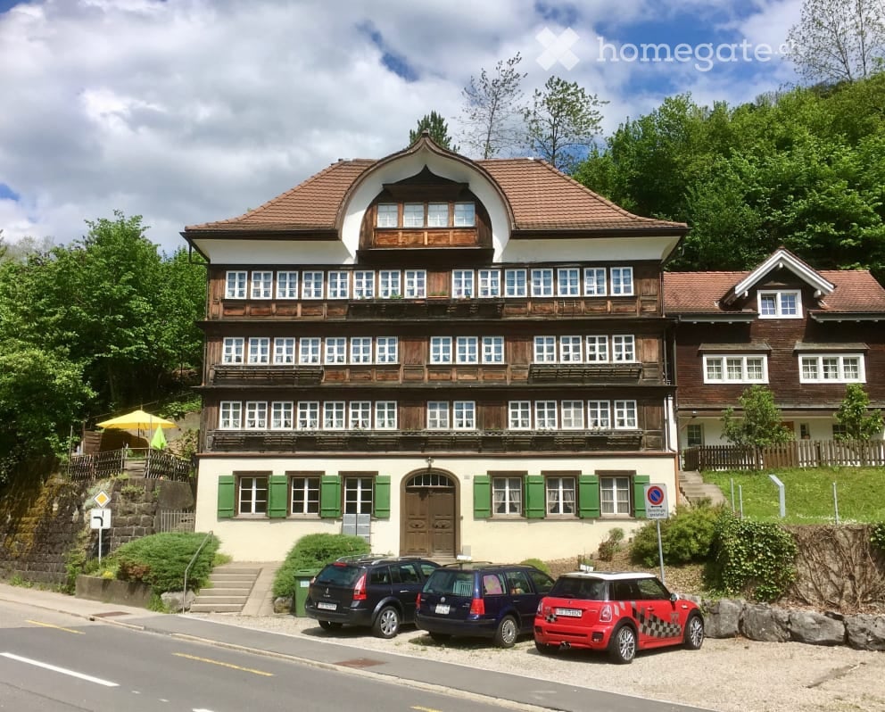 A three-story traditional house with a wooden facade and green shutters, featuring multiple windows and a central entrance. The building is surrounded by greenery and has several parked cars in front.