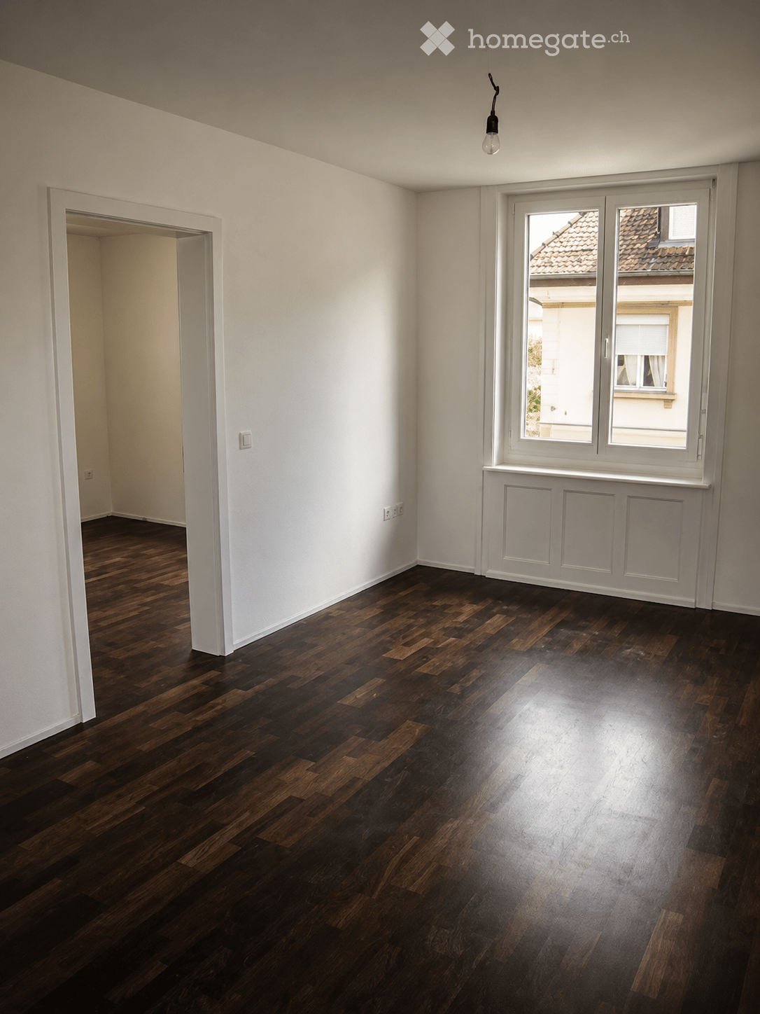 Empty room with dark wooden flooring, white walls, and large windows showing a view of neighboring buildings.