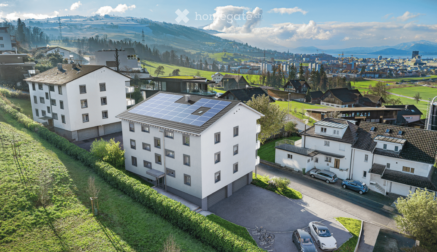 Aerial view of a residential area with multiple houses, solar panels on one roof, parked cars, and a scenic valley view in the background.
