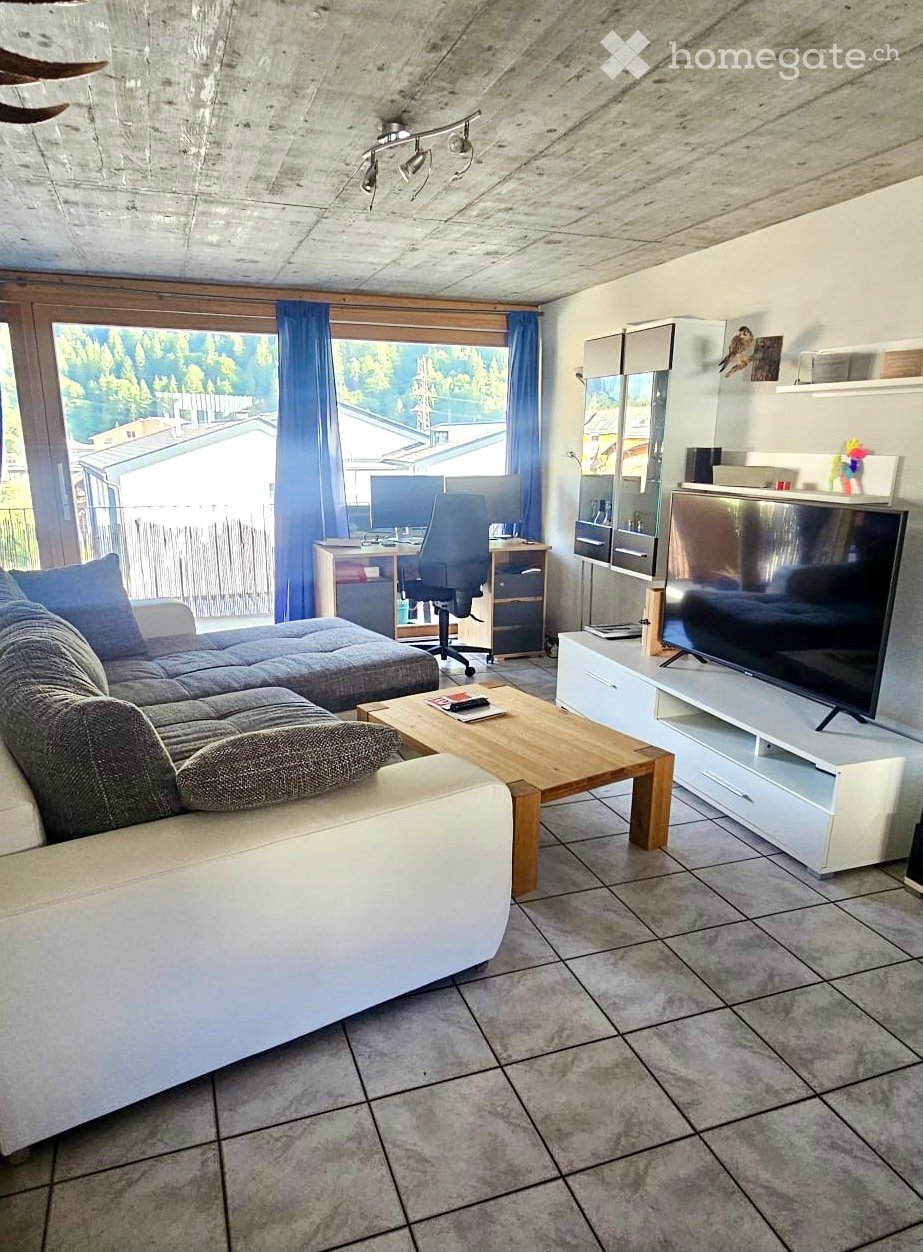 Modern living room with a grey sectional sofa, wooden coffee table, flat-screen TV on a white stand, desk with chair, and large windows with blue curtains.