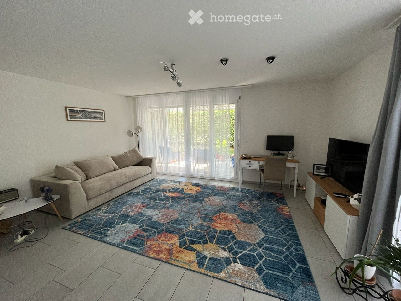 Bright living room with a beige sofa, colorful geometric rug, coffee table, desk with chair, television, and floor lamp. Large windows with sheer curtains let in natural light.