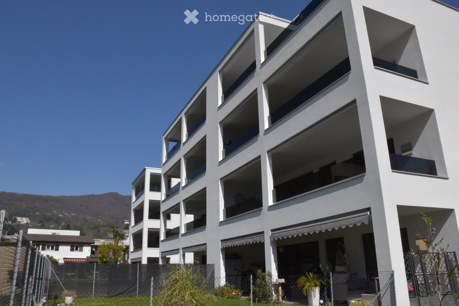 Modern multi-story residential building with white facade, large windows, and balconies. The building features contemporary design with clean lines and geometric shapes.