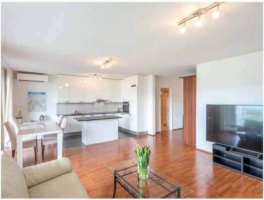 Modern, open-plan living and dining area with light wood flooring, white kitchen cabinets, a central island, a sofa, coffee table, dining table with chairs, and a television on a stand.