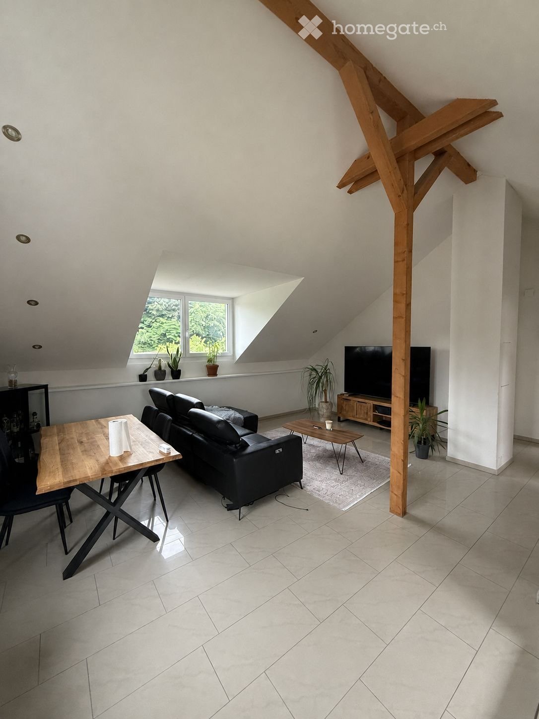 Modern living room with a black leather sofa, wooden dining table, chairs, TV stand, and potted plants. The room features exposed wooden beams and large windows with a view of greenery.