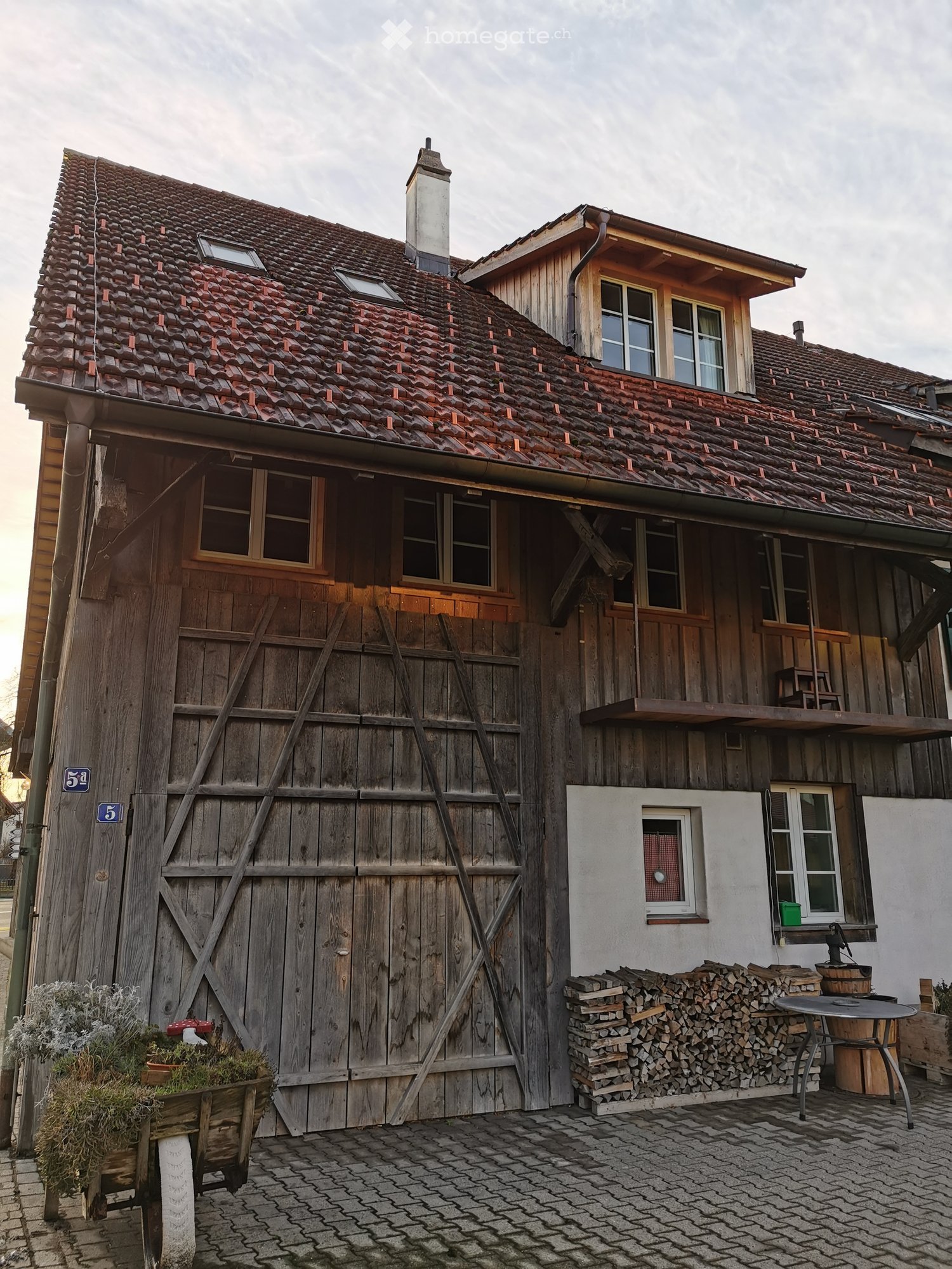 Traditional wooden house with a red-tiled roof, multiple windows, and a wooden garage door. A stack of firewood and a small table are visible in the foreground.