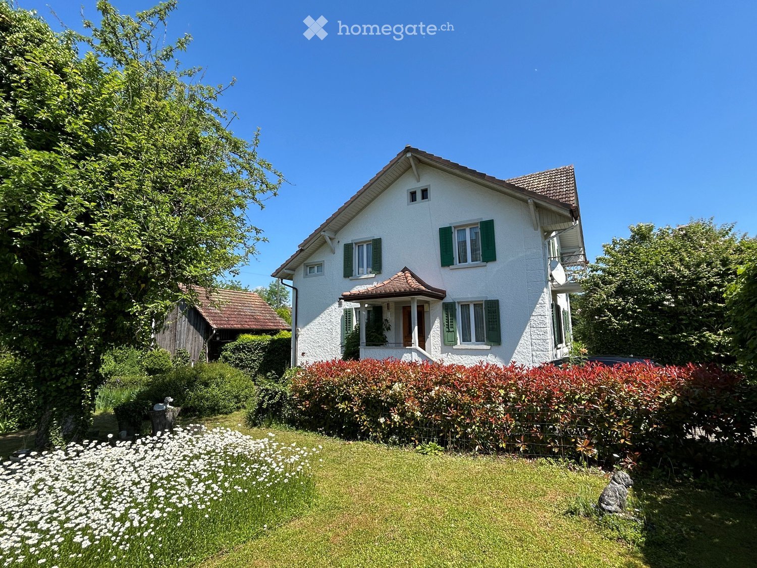 Two-story white house with green shutters, red-tiled roof, and a small balcony on the right side. The house is surrounded by a well-maintained garden with a hedge, flowers, and trees.