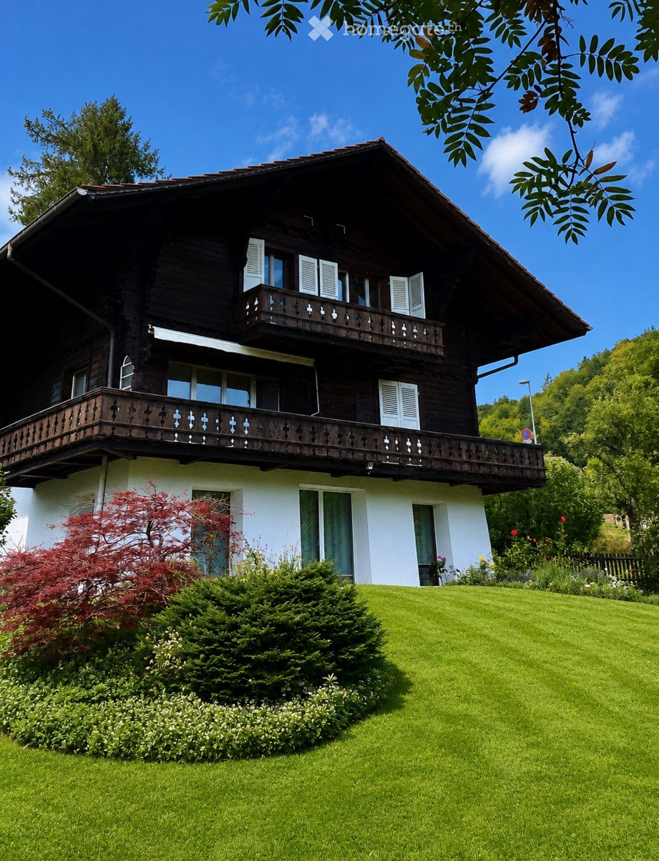 Two-story wooden house with white shutters, balconies, and a well-maintained garden with green lawn and shrubs.