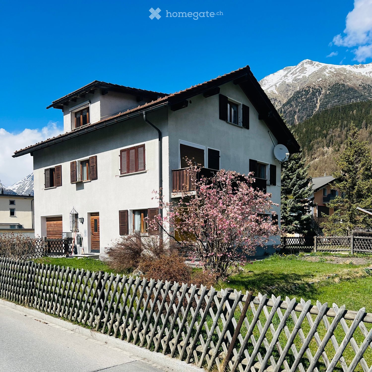 Two-story white house with brown shutters, wooden door, balcony, and surrounding garden with blooming trees and mountains in the background.