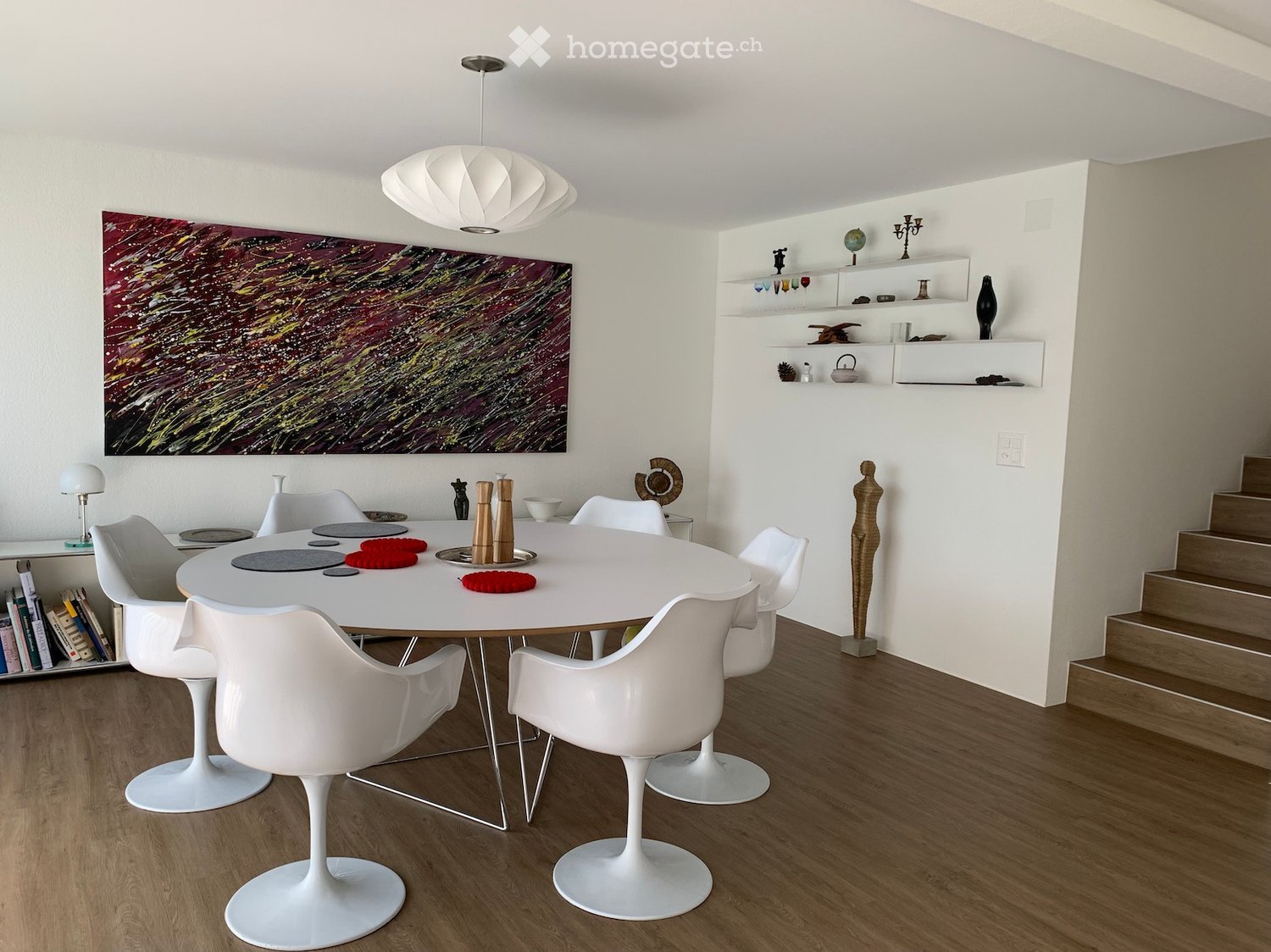 Modern dining room with a white round table, white chairs, shelves with decorative items, a large abstract painting, and a staircase leading upwards.
