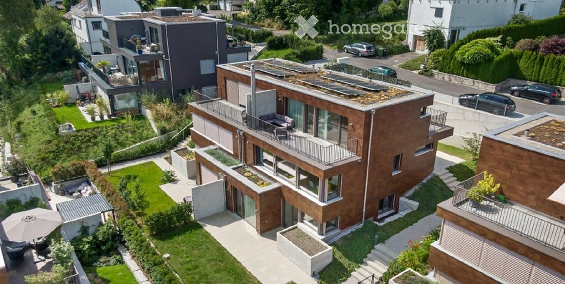Aerial view of a modern, multi-story residential building with a green roof, balconies, and surrounding landscaped gardens. The building features contemporary architecture with large windows and a mix of wood and concrete materials.