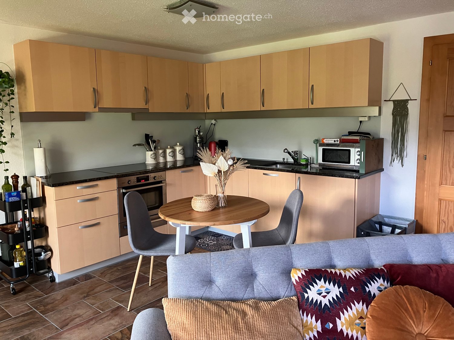 A modern, open-plan living and dining area with a grey sofa, patterned cushions, a round wooden dining table with two chairs, a kitchen area with light wood cabinets, an oven, microwave, and a sink.