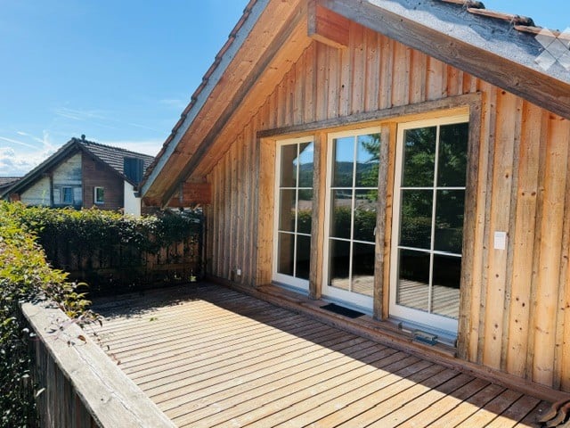 Wooden balcony with large glass doors leading to a wooden deck, overlooking a scenic view of trees and a neighboring house.