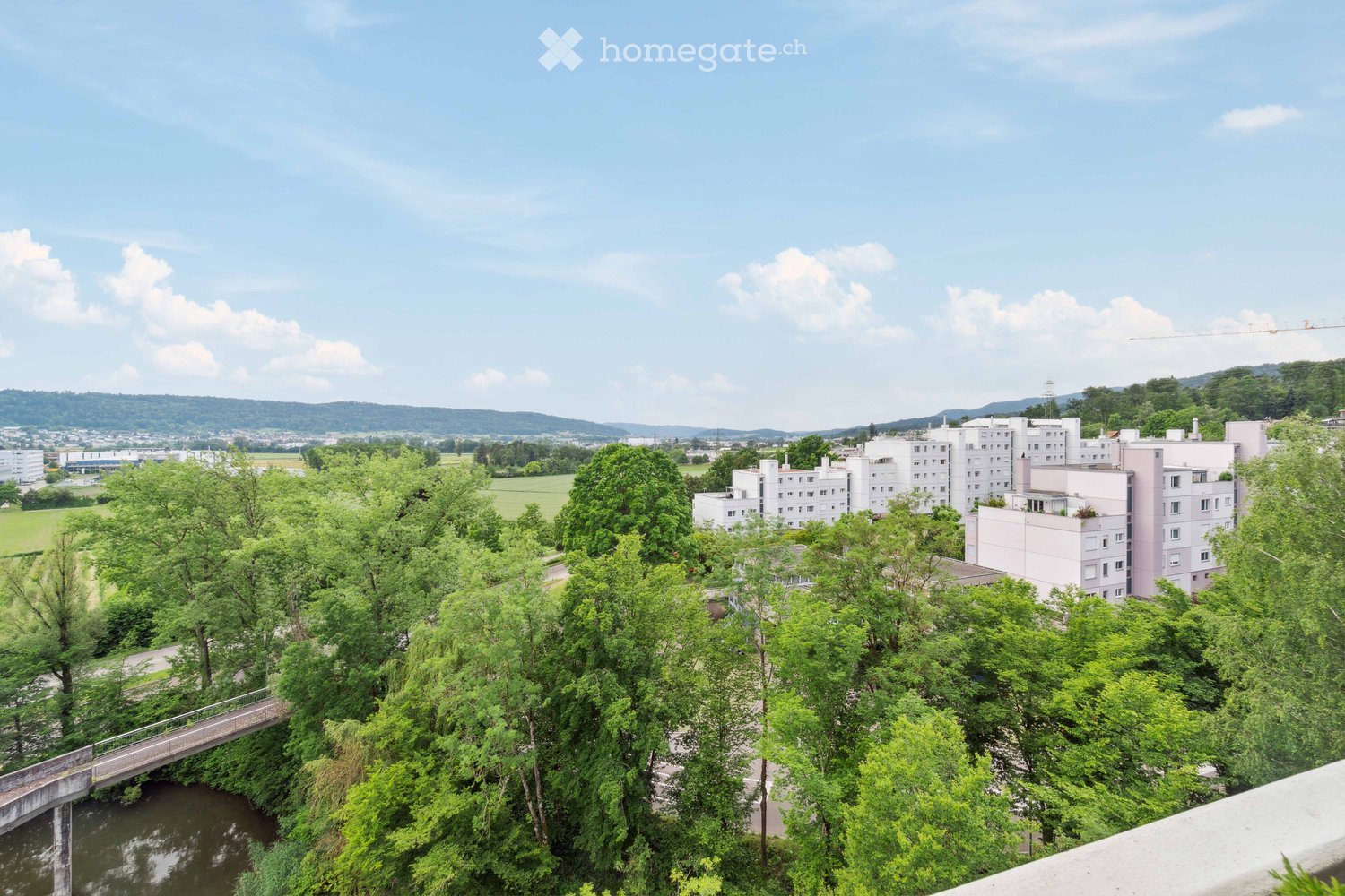 A panoramic view of a residential area with multiple buildings, surrounded by lush green trees and a river running through the foreground.